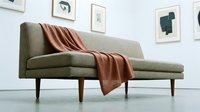 Beige mid-century modern sofa with wooden legs and rust-colored throw blanket in minimalist living room interior design setting.