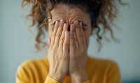 Woman with curly hair wearing yellow sweater covering face with hands expressing emotional distress, anxiety, shame or crying against light background.