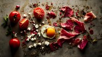 Fresh ingredients scattered on dark background with sunny side up egg, prosciutto, tomatoes, and feta cheese crumbles creating rustic food composition.