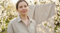 Young Caucasian woman in linen shirt hanging laundry outdoors among blooming spring trees with natural sunlight creating warm domestic lifestyle scene.
