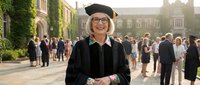 Smiling mature Caucasian woman in graduation cap and gown celebrating academic achievement at university campus ceremony with crowd of graduates in background.