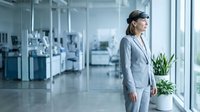 Woman in business attire standing by window in modern laboratory or medical facility with scientific equipment and plants in background.