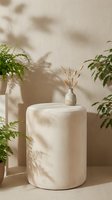 Minimalist white ceramic side table with dried grass vase surrounded by green plants casting natural shadows on neutral wall in modern interior design.