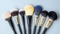 Professional makeup brushes arranged in fan shape on gray background, featuring soft synthetic bristles in various sizes for cosmetic application and beauty artistry.