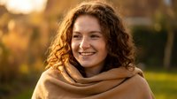 Young woman with curly hair wrapped in cozy blanket smiling warmly during golden hour sunset outdoors in autumn park for lifestyle and wellness content.