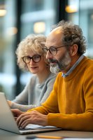 Senior Caucasian couple working together on laptop computer, man in yellow sweater and woman with curly hair wearing glasses in modern office setting.