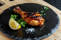 Grilled chicken leg with honey glaze served on black slate plate, garnished with fresh parsley and lemon wedge, close up view of gourmet meal presentation.