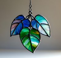 Stained glass leaves pendant in vibrant blue and green tones with metallic frame, illuminated from behind on neutral background. Decorative suncatcher art.
