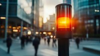 Glowing red traffic light pole against blurred urban cityscape at sunset with silhouettes of people walking on street and modern glass buildings in background.
