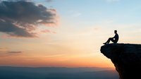 Silhouette of person sitting on cliff edge against dramatic sunset sky and mountain landscape. Concept of solitude, achievement and contemplation.