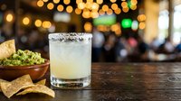 Margarita cocktail with salt rim served on bar counter with tortilla chips and guacamole dip, blurred festive lights in background, perfect for restaurant and beverage marketing.