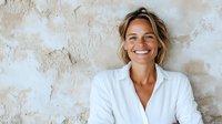 Cheerful blonde woman in white shirt smiling against textured wall background. Natural daylight creates soft shadows highlighting genuine happiness and positive lifestyle.