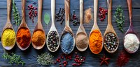 Colorful spices and herbs in wooden spoons arranged in row on dark background with scattered peppercorns, berries and rosemary sprigs.