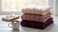 Cozy autumn scene with stacked knit sweaters in warm tones, steaming mug, eyeglasses and open book by bright window for fall lifestyle and comfort themes.