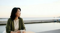 Young East Asian woman in olive cardigan gazing at calm waterfront at dusk, contemplative mood for lifestyle and wellness content.