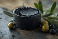 Black charcoal soap bar with green and black olives on dark wooden surface. Natural organic skincare product with olive leaves creates spa atmosphere.