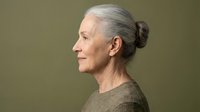 Senior woman with gray hair in bun, fair complexion, wearing olive sweater, smiling in profile against neutral background. Portrait for aging, wellness, lifestyle content.