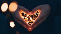 Hands forming a heart shape holding glowing fairy lights against dark bokeh background, symbolizing love and romance.