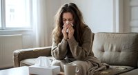Young woman with long brown hair blowing nose into tissue while sitting on couch at home, feeling unwell with cold or flu symptoms during winter season.