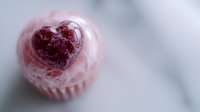 Pink frosted cupcake with heart-shaped chocolate decoration on top, photographed from above on marble surface, soft focus creates romantic mood for Valentine's Day treats.