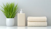 Minimalist bathroom essentials with cream-colored towels, soap dispenser, and decorative grass plant in white pot against light gray background for spa and wellness design.
