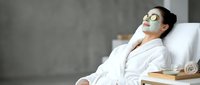 Young Asian woman relaxing in white robe with green clay face mask and cucumber slices on eyes at spa wellness center.