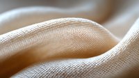 Soft beige linen fabric with gentle folds and waves creating abstract texture pattern. Natural cotton material macro photography shows detailed textile weave structure.