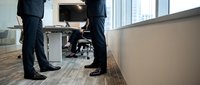 Businessmen in formal suits shake hands in modern office corridor with colleague in wheelchair, representing workplace inclusion and professional partnership.