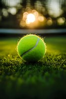 Tennis ball on green grass court at sunset with golden bokeh lights in background, close-up macro photography showing detailed texture and warm atmosphere.