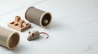 Minimalist eco-friendly cat toys on marble surface, including wooden puzzle, felt mouse, and cardboard tunnels in neutral tones.