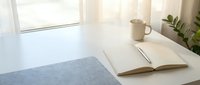 Bright workspace with open notebook, pen, coffee mug and potted plant on white desk near window with natural light, ready for creative work or study.