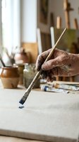 Aged artist hand with paint-stained fingers holding a brush, applying blue paint to canvas in a rustic studio with easels and jars.