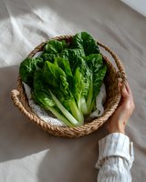 Fresh green bok choy in woven basket held by hand wearing white sleeve against neutral linen background, natural light food photography styling.