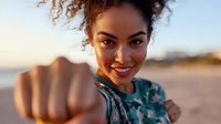 Young African American woman with curly hair smiling and playfully extending fist toward camera on sunny beach at golden hour, wearing teal patterned top.