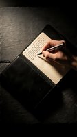 Person with medium skin tone writing handwritten notes in open leather journal with pen on dark textured surface, suitable for education, creativity, journaling, and business content.