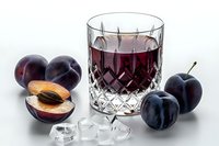 Crystal glass of fresh plum juice surrounded by whole and sliced ripe plums and ice cubes on white background, perfect for beverage advertising.