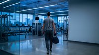 Athletic man walking through modern gym with sports bag during evening workout session in contemporary fitness center with city skyline view.