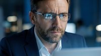 Professional businessman with glasses analyzing data on laptop screen in modern office environment with blue lighting and focused concentration.