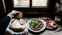 Rustic breakfast spread with sourdough bread, fresh salad, prosciutto, and soft cheese by window. Natural light creates moody atmosphere on wooden table.