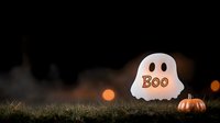 Glowing ghost decoration with boo text and small pumpkin on dark grass with bokeh lights in background creates spooky Halloween atmosphere.