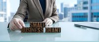 Professional businessman arranging wooden letter blocks spelling small business brand on modern office desk with city skyline background.
