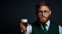 Young caucasian male with red beard wearing glasses, green vest and tie holds craft beer glass with foam head against dark background, looking at camera with slight smile.
