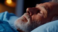 Senior Caucasian man with gray beard sleeping peacefully on blue pillow in warm ambient lighting, side profile view showing relaxed facial expression.