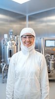 Smiling female technician in white cleanroom suit and safety glasses standing in modern pharmaceutical manufacturing facility with industrial equipment.