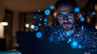 Caucasian male professional with glasses working on laptop at night surrounded by glowing blue digital particles and technology visualization effects.
