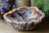 Natural agate geode crystal bowl with orange and gray banded layers, rough outer surface and polished interior cavity, displayed on wooden surface with flowers.