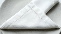 Folded white linen napkin with decorative hemstitching detail on neutral beige surface, ready for table setting, event styling, hospitality design.