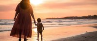 Mother and toddler walking hand in hand on beach at sunset with golden light reflecting on wet sand and gentle waves in background.