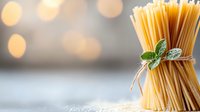 Bundle of raw spaghetti pasta tied with twine and fresh basil leaf against soft bokeh background, showcasing Italian cuisine ingredients.