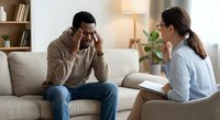 Middle-aged Hispanic man experiencing headache during therapy session with female psychologist in modern office setting for mental health treatment.